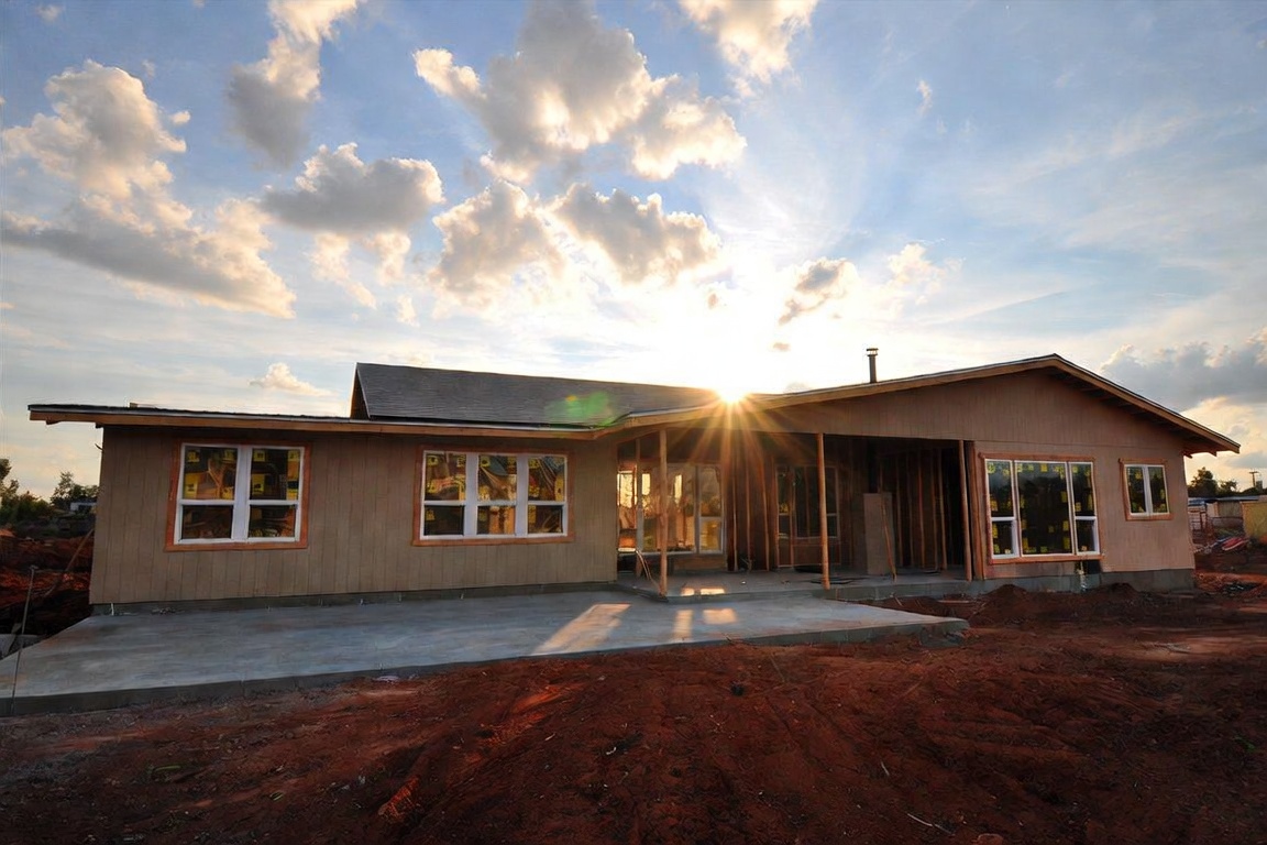 Single-story house under construction on a gray concrete slab foundation, surrounded by red Oklahoma clay soil at sunset, with an unfinished porch and no basement.